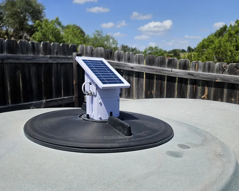 A Trivertix monitor with its solar panel, mounted in the lid of a poly water tank under a blue sky