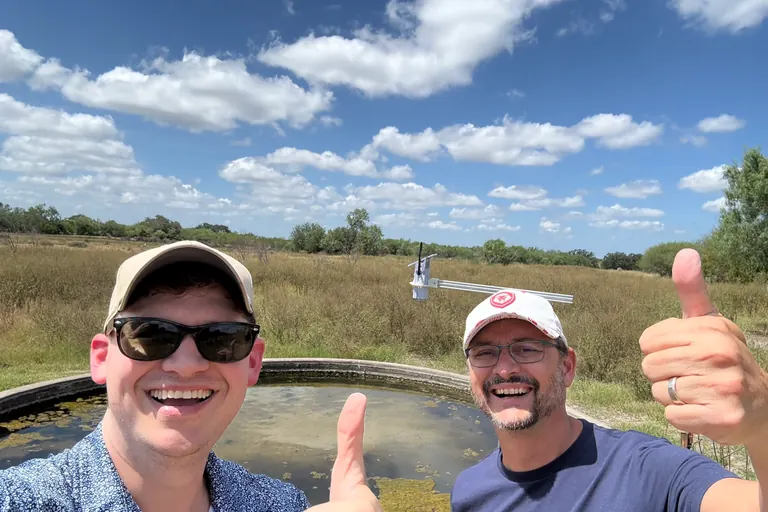 Two people give a thumbs up beside a concrete stock tank, with a Trivertix monitor on a pole behind them